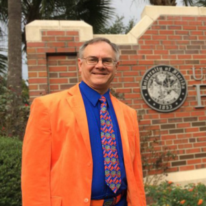 Larry Kubiak is standing outside a University of Florida campus entrance. He is wearing an orange suit with a blue shirt and a Gators tie while smiling.