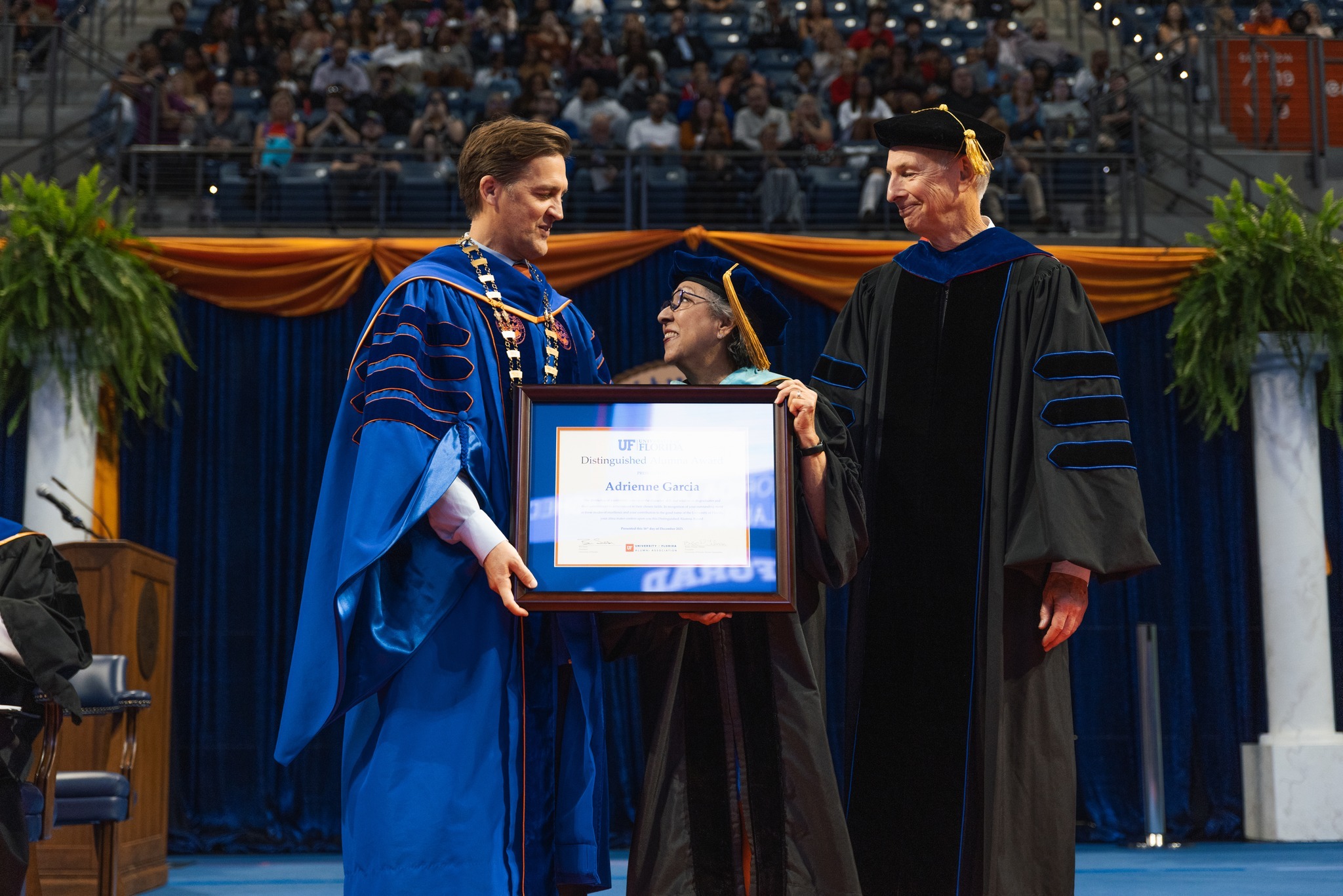 This is an image from the fall doctoral commencement ceremony. The photo captures a moment when Adrienne Garcia, in academic regalia, is receiving the UF Distinguished Alumni award. UF President Ben Sasse and UF College of Education Dean Glenn Good are on either side of her on the stage, also in academic regalia. The background shows an audience seated, watching the ceremony. The stage is decorated with orange and blue colors.
