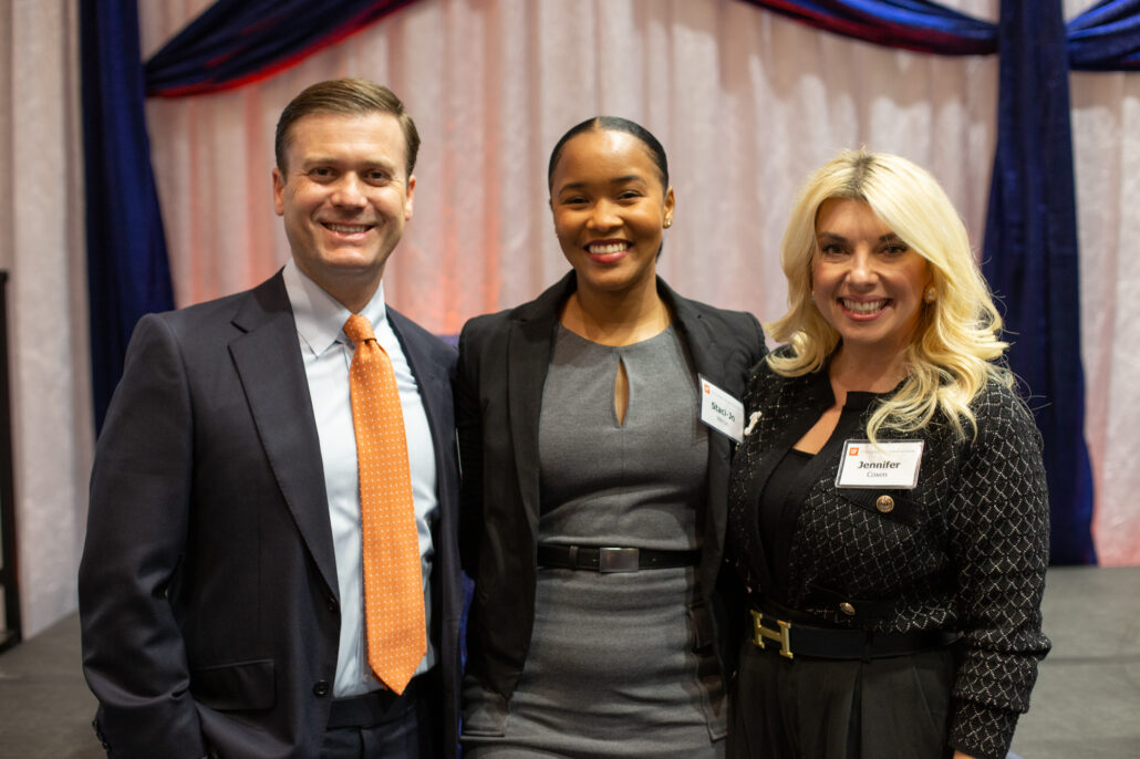 Three people stand together in front of a draped background at the annual UF College of Education Scholarship and Awards dinner. They are all dressed in formal attire, and two have name tags. The person on the left wears a dark suit with an orange tie. The student in the middle is dressed in a grey dress and black blazer. Alumna and donor Jennifer Coxen, on the right, wears a patterned black dress and has blonde hair.