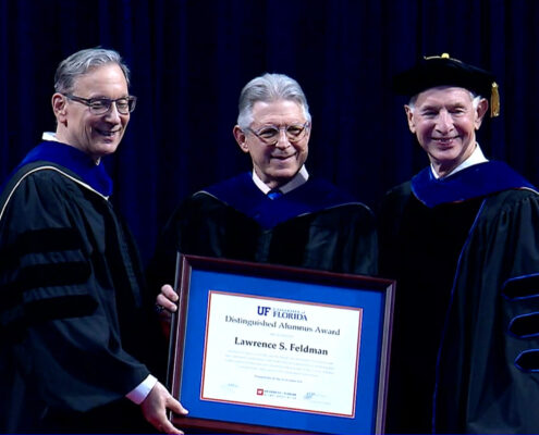 Three individuals in academic regalia on a dark stage hold a framed University of Florida Distinguished Alumnus Award for Lawrence S. Feldman.