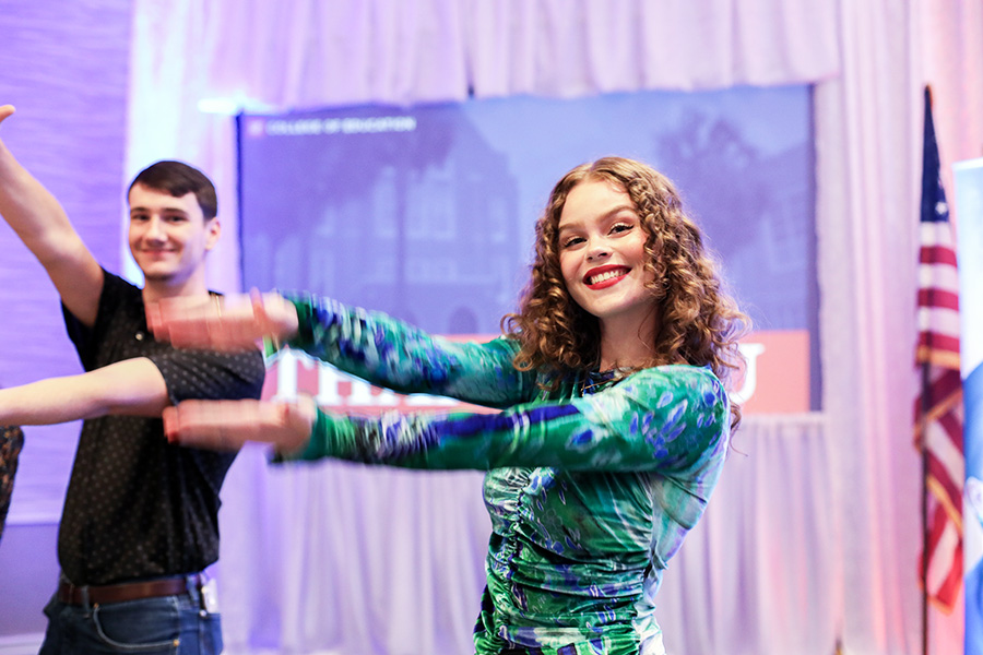 At an indoor College of Education event, two participants perform the Gator Chomp by extending their arms forward, right arm over left, and mimicking an alligator’s mouth opening and closing; the person in the foreground is in a green patterned top, the other participant is to the left, and a U.S. flag and event banner are in the background.
