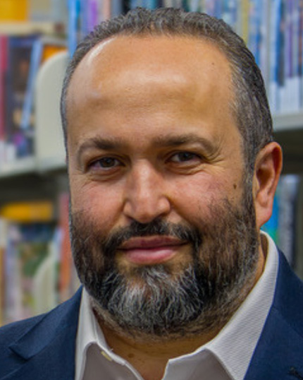 A person with a dark beard, speckled with grey, wearing a navy blazer and white collared shirt standing in front of library bookshelves.