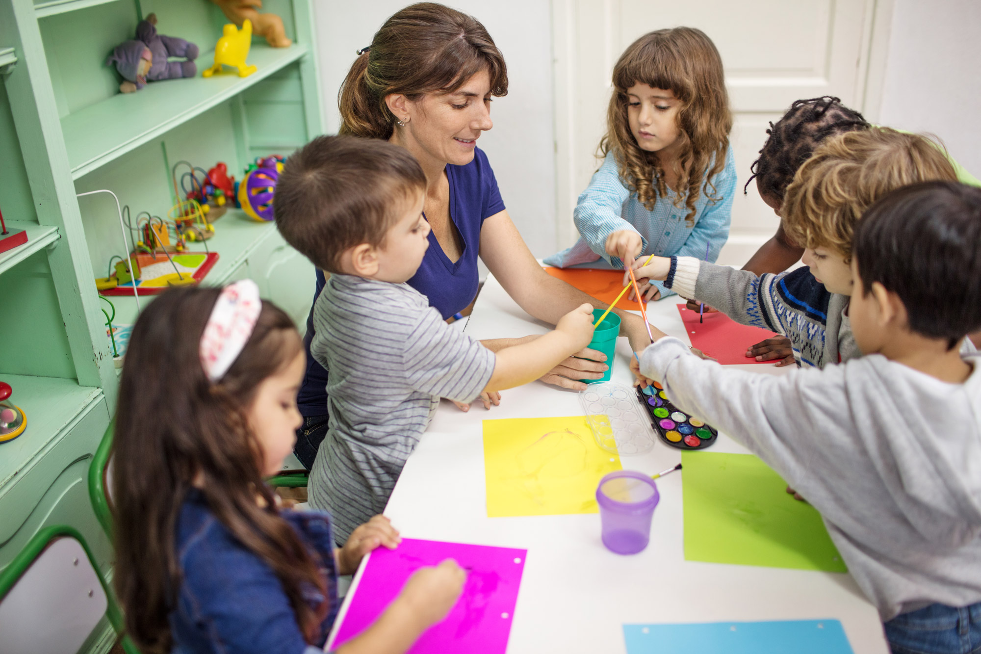 Preschool kids with teacher painting at desk in kindergarten. Teacher sitting with students coloring in classroom.