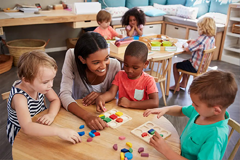 Online_early_childhood_program Preschool classroom scene: a teacher helps four young children sort colorful geometric blocks and a wooden puzzle at a round table, while other children work at another table in a bright, cozy playroom.