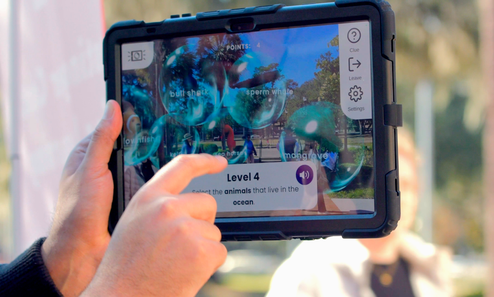 Hands hold a tablet outdoors, tapping an educational AR game screen that reads “Level 4: Select the animals that live in the ocean,” with floating bubbles labeled bull shark, sperm whale, clownfish, seahorse, and mangrove.