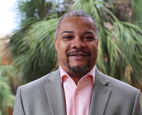 A headshot of Rob Moore. He is wearing a grey blazer and standing in front of a palm tree.