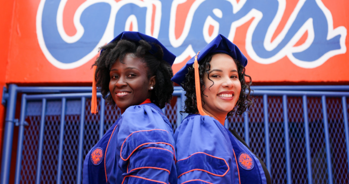A posed photograph of Bertrhude Albert, Ph.D. (left) and Priscilla Zelaya, Ph.D. (right) in UF doctoral regalia in the football stadium on campus. They are both smiling.