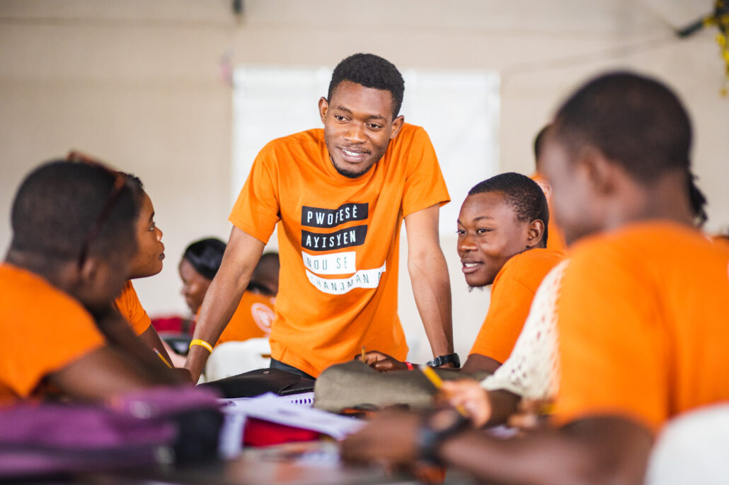 A Haitian teacher in an orange shirt with text in Haitian Creole. They are leaning over a desk and talking to other teachers who are also wearing orange shirts.