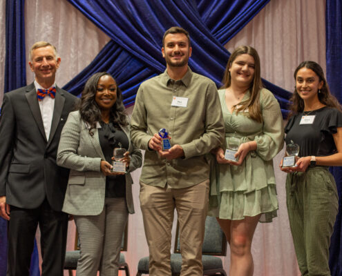 UF College of Education Dean Glenn Good posing with the 2022 student winners at the 19th annual Scholarship and Awards dinner. From left to right are Yasmeen Merriweather, Max Sommer, Nicole Wasserman and Grace Miles. All of them are in business casual attire and are standing against a white and blue draped fabric background.