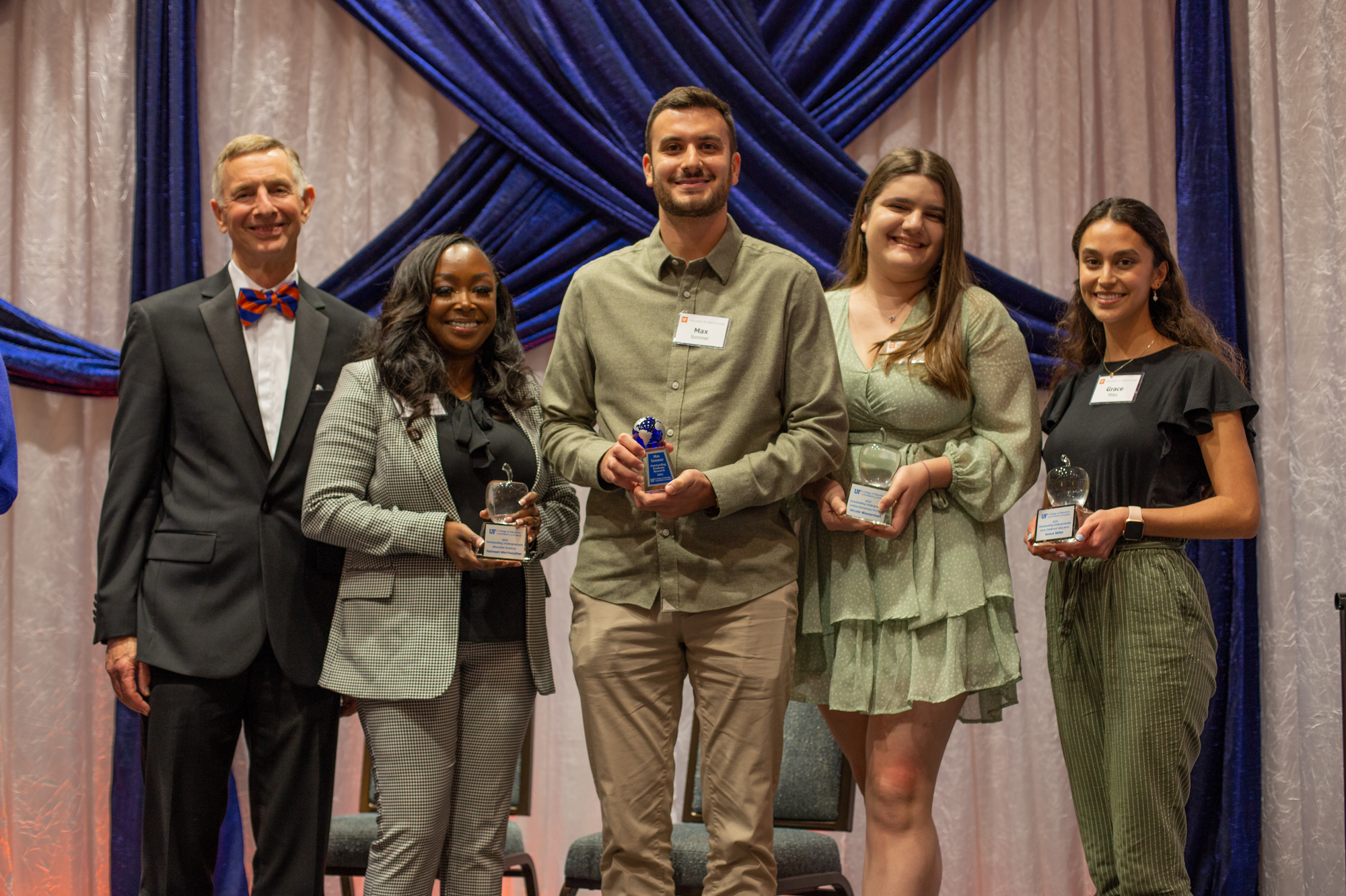 UF College of Education Dean Glenn Good posing with the 2022 student winners at the 19th annual Scholarship and Awards dinner. From left to right are Yasmeen Merriweather, Max Sommer, Nicole Wasserman and Grace Miles. All of them are in business casual attire and are standing against a white and blue draped fabric background.
