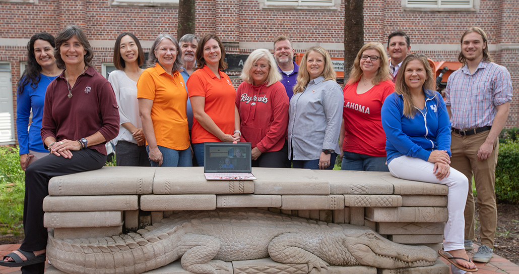 A group of people in SEC school shirts are posing in the Dorfeld Plaza at Norman Hall. They are posing around the gator book bench, and people are on a laptop screen.