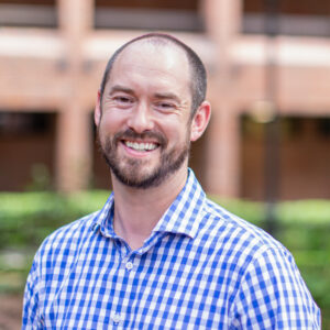 A headshot of Christopher Thomas. He is wearing a blue checkered shirt and is standing in the Norman Hall Courtyard at the University of Florida College of Education.