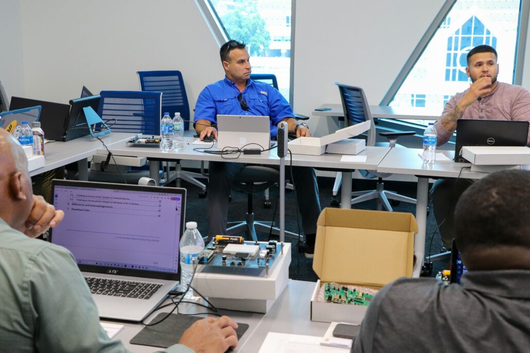 A close-up of a conference room setting where several individuals are seated at tables with laptops and electronic devices. One person in a blue shirt and sunglasses is working on a laptop, while another in a pink shirt sits nearby with a laptop. An open box with electronic components is visible in the foreground. The room is bright with large windows providing an outside view. Portions of this alt text were created or edited using generative AI.