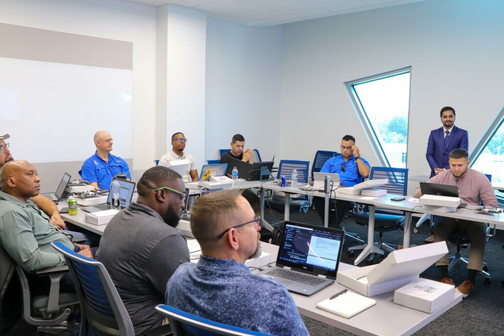 A group of about nine people in a modern conference room engaged in a meeting. They are seated at tables arranged in a U-shape, with laptops, notebooks, and various electronic devices in front of them. The people seated are all dressed casually. A person in a blue suit stands near the windows, addressing the group. The room has large windows letting in natural light. Portions of this alt text were created or edited using generative AI.