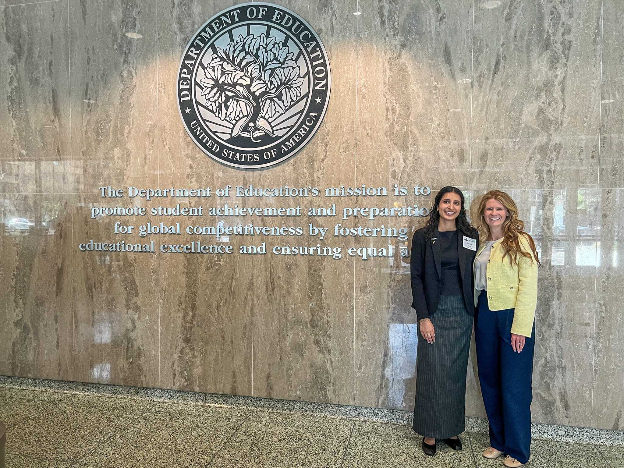 Two women posing in front of a wall with text and a logo in the Capitol Hill building.