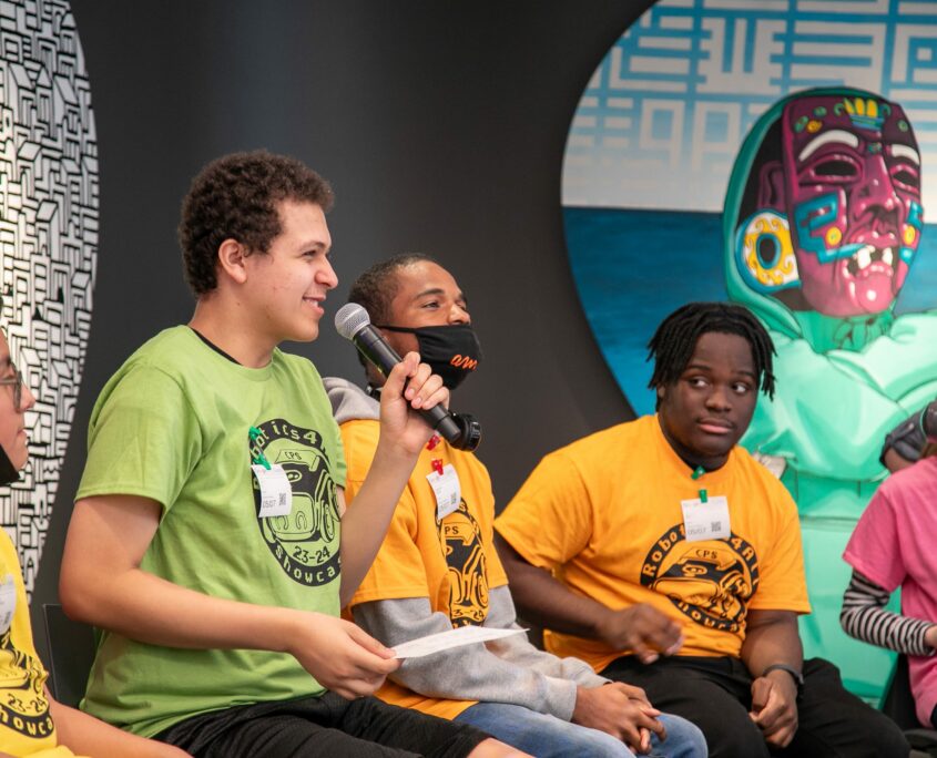 Several young students are seated on a stage with a backdrop featuring a large screen displaying a computer chip-inspired graphic; they are attentively participating in a discussion, with one student speaking into a microphone.