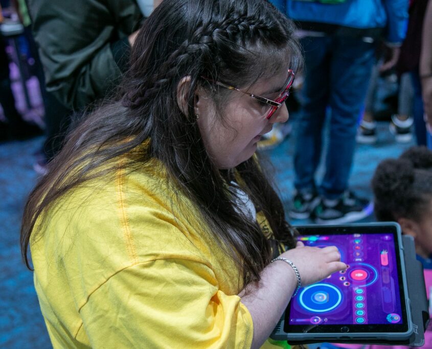 A young woman is seated, focusing intently on a tablet displaying colorful, interactive programming interfaces, surrounded by a bustling event environment.