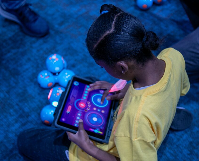 A young girl is seated on the floor interacting with a bright, colorful display on a tablet amidst several illuminated robotic devices on the carpeted floor.