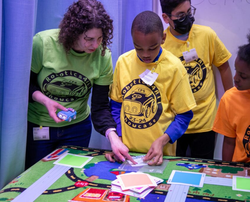 Students gather around a colorful educational mat on a table, interacting with various learning materials and tools during an educational event.