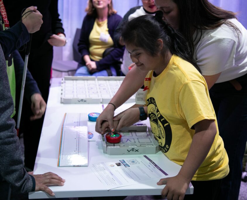 A young student in a yellow shirt engages with interactive educational technology at a table surrounded by adults observing and assisting.