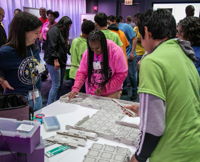 A group of students and adults focus on assembling or manipulating modular educational components on a white table during a robotics demonstration or workshop.