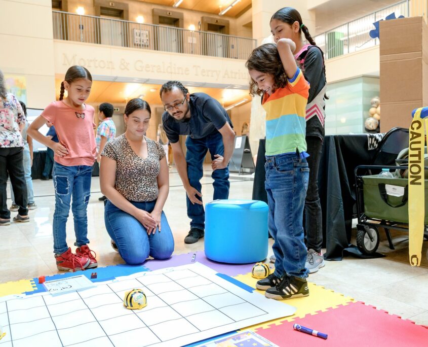 Children and adults are engaged in a public event at a spacious atrium, centered around an educational display on the floor. A colorful mat with grid lines features programmable robots, where children observe and interact with the setup. Family members are nearby, watching the activities.