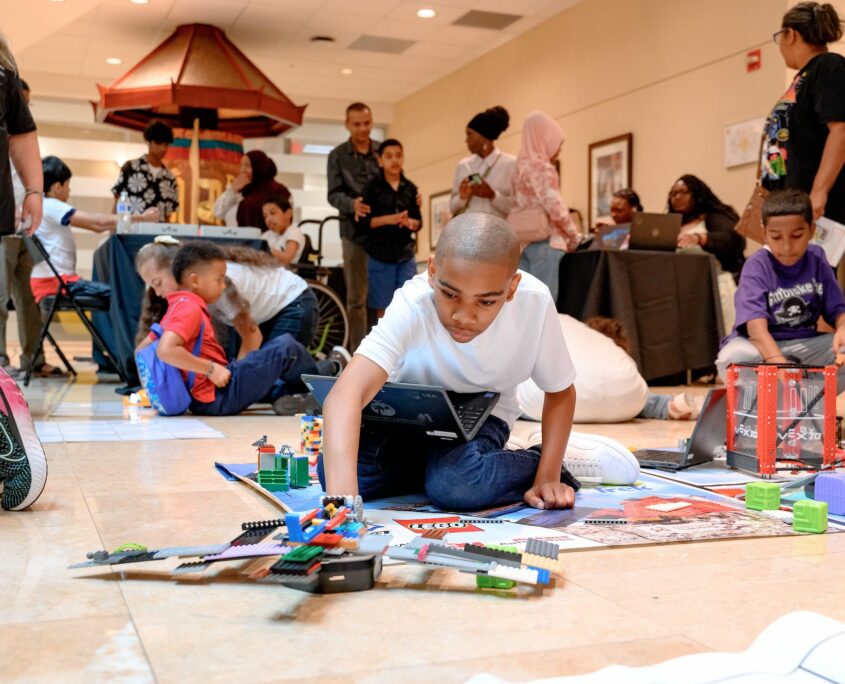 A young child is focused on programming on a laptop in a busy indoor setting, surrounded by building toys and robotics kits. Other children and adults are attending the event in the background.