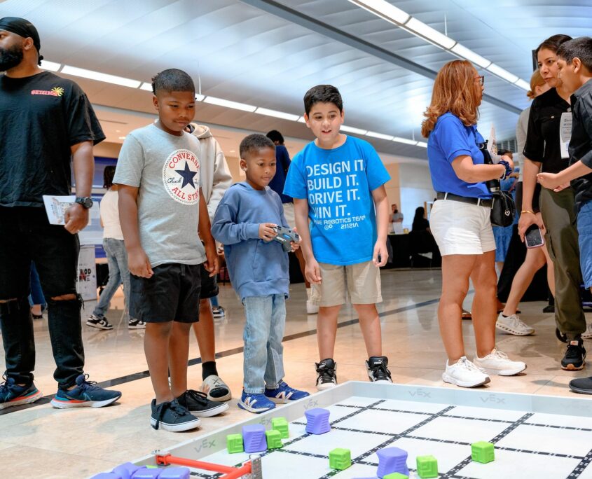 A group of people, including children and adults, engage around a robotics grid display at an indoor event. The scene shows interactive participation with a focus on education and technology activities.