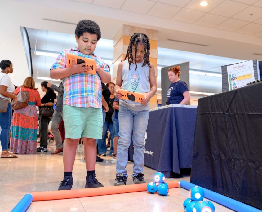 Two children use remote control devices to maneuver robotic balls on the floor during a robotics event, showcasing the integration of technology and education. A busy environment surrounds them with more participants and displays in the background.