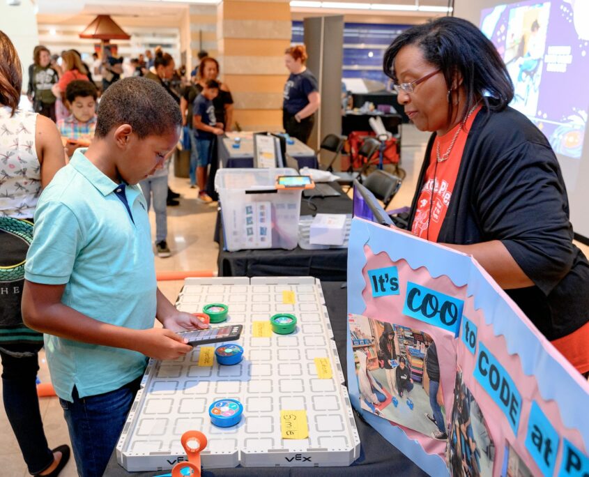 Children use interactive coding tools on a display table at a technology event, where participants engage in digital learning activities, surrounded by a bustling crowd.