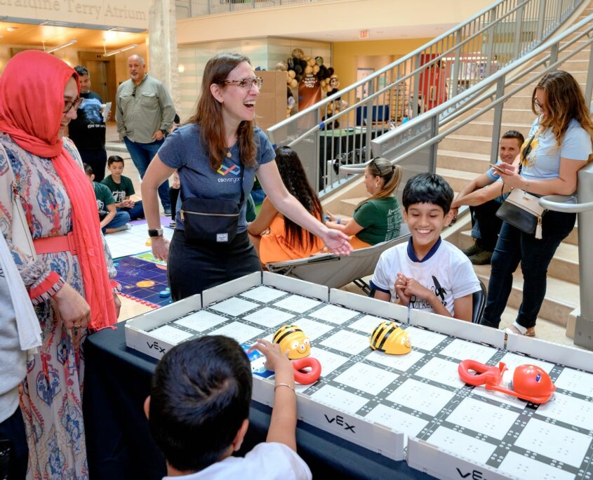 Children use interactive coding tools on a display table at a technology event, where participants engage in digital learning activities, surrounded by a bustling crowd.