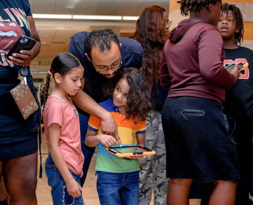 At a busy educational event, a child interacts with a tablet while a parent helps over their shoulder, surrounded by other attendees engaged in various activities.