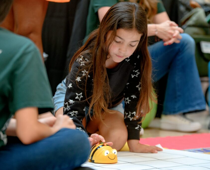 A young girl interacts with a robotics toy modeled after a bee with a yellow body and black stripes while sitting on the floor.