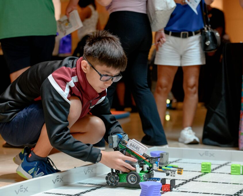 A boy maneuvers a robotics toy with wheels. The toy is being placed on a mat with grid marks amongst other toys that look like green cubes.