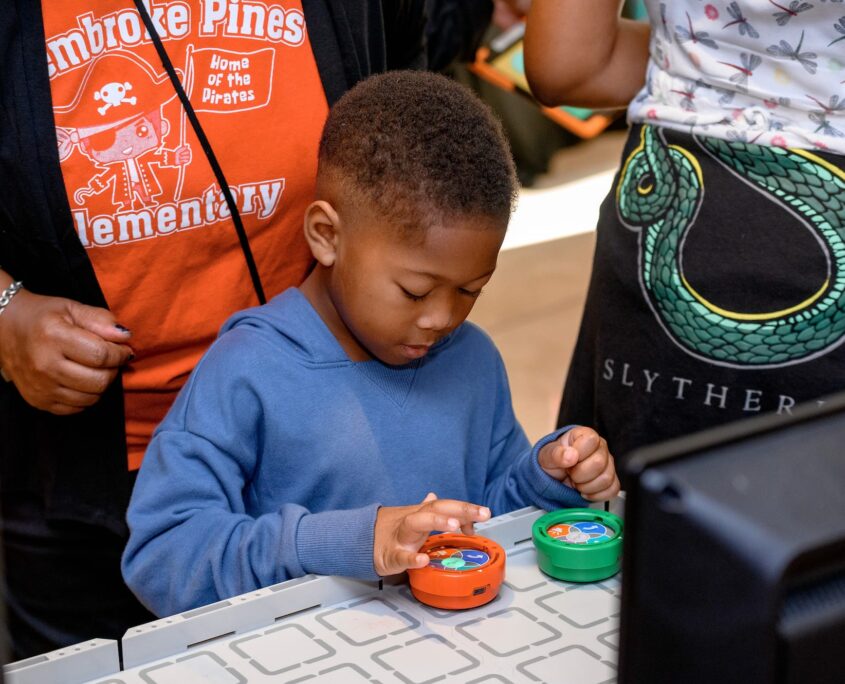 A young boy interacts with a circular, orange robotics toy on a table next to a circular green robotics toy.