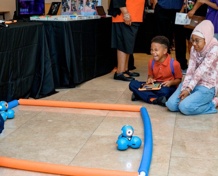 Two students excitedly interact with a tablet while looking at two robotics devices on the tile floor that appear to consist of blue balls clumped together.