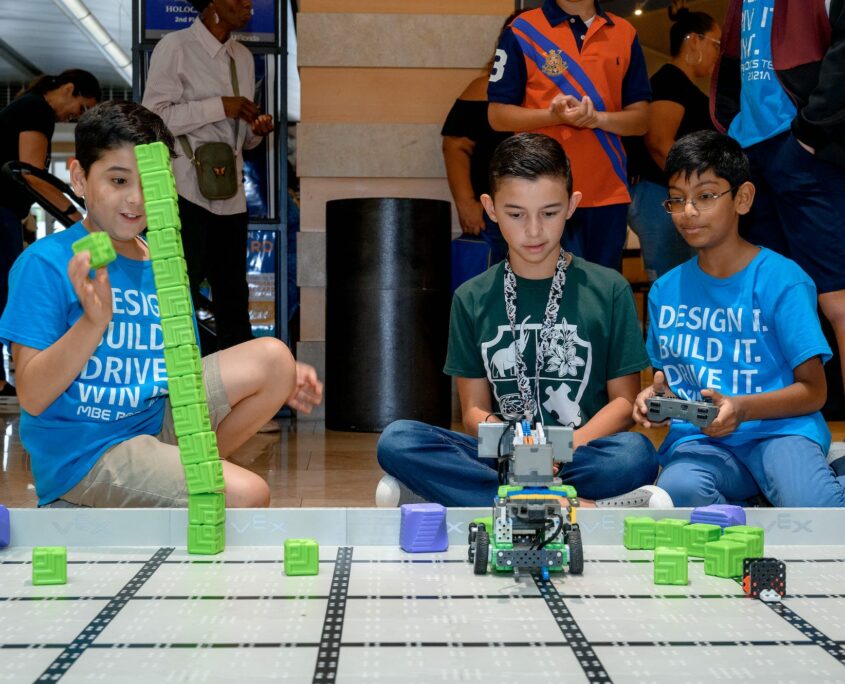 Children sit on the floor engaged with a robotics activity, controlling and building robots on a grid mat that is littered with blocks and components, in an indoor public space bustling with other attendees.