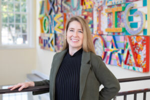 Avery Closser wearing a blazer and smiling in front of a colorful art installation featuring letters of the alphabet