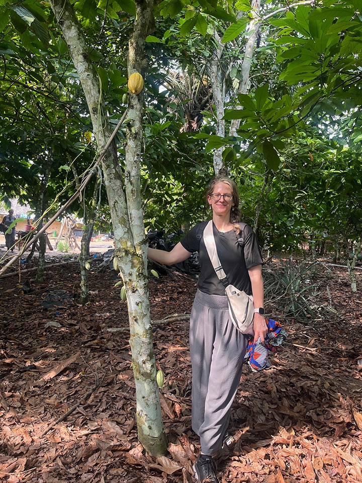 A person stands next to a tree in a lush forest environment, holding onto its trunk. They are dressed casually in a black t-shirt, grey pants, and black shoes, and are carrying a light-colored crossbody bag and some patterned cloth in their left hand. The ground is covered with leaves, and there are multiple trees and greenery in the background.