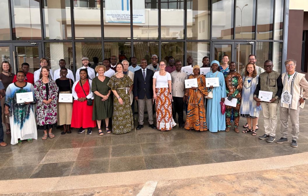 Approximately thirty adults pose for a group photo in front of a building with large glass doors; several hold certificates, and the group wears a mix of formal attire and brightly patterned traditional clothing on a tiled entryway.