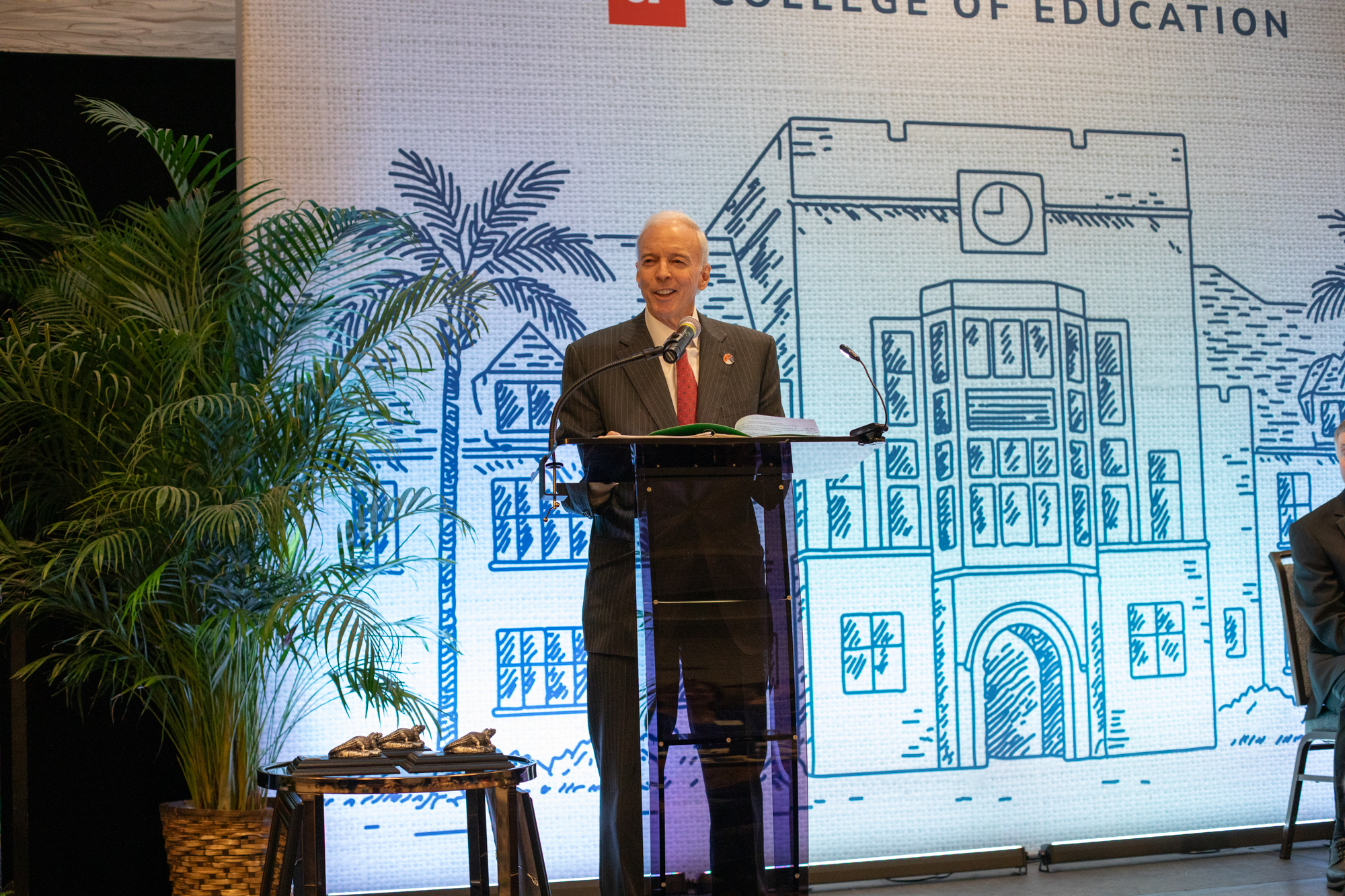 Speaker in a dark pinstripe suit and red tie stands at a clear lectern with microphones, reading from a green folder, on a stage with potted palms and a “College of Education” backdrop showing a sketched building with a clock.