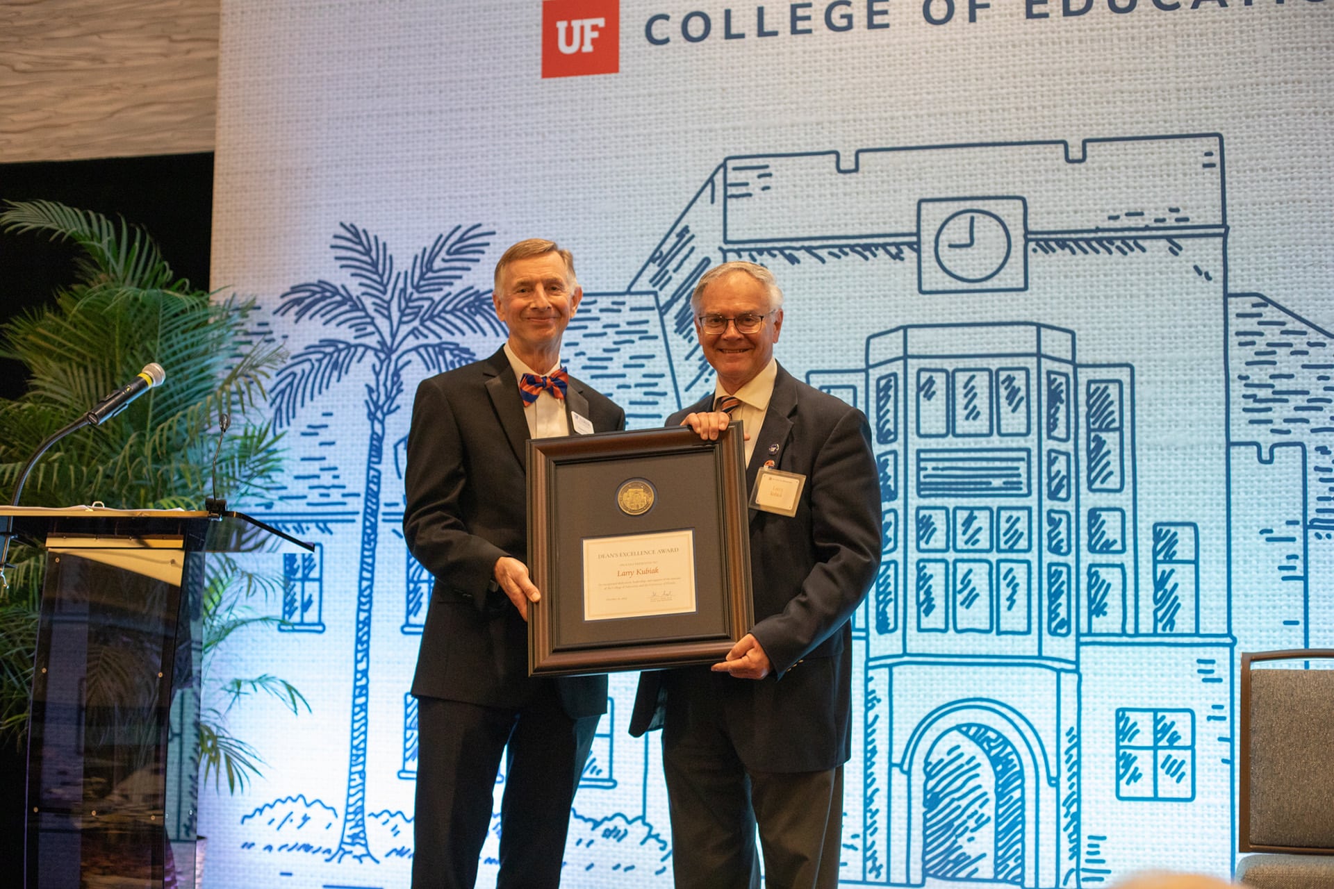 Two people in suits hold a large framed award on stage in front of a UF College of Education banner and a blue line-art building backdrop, with a podium and palm plants to the left.