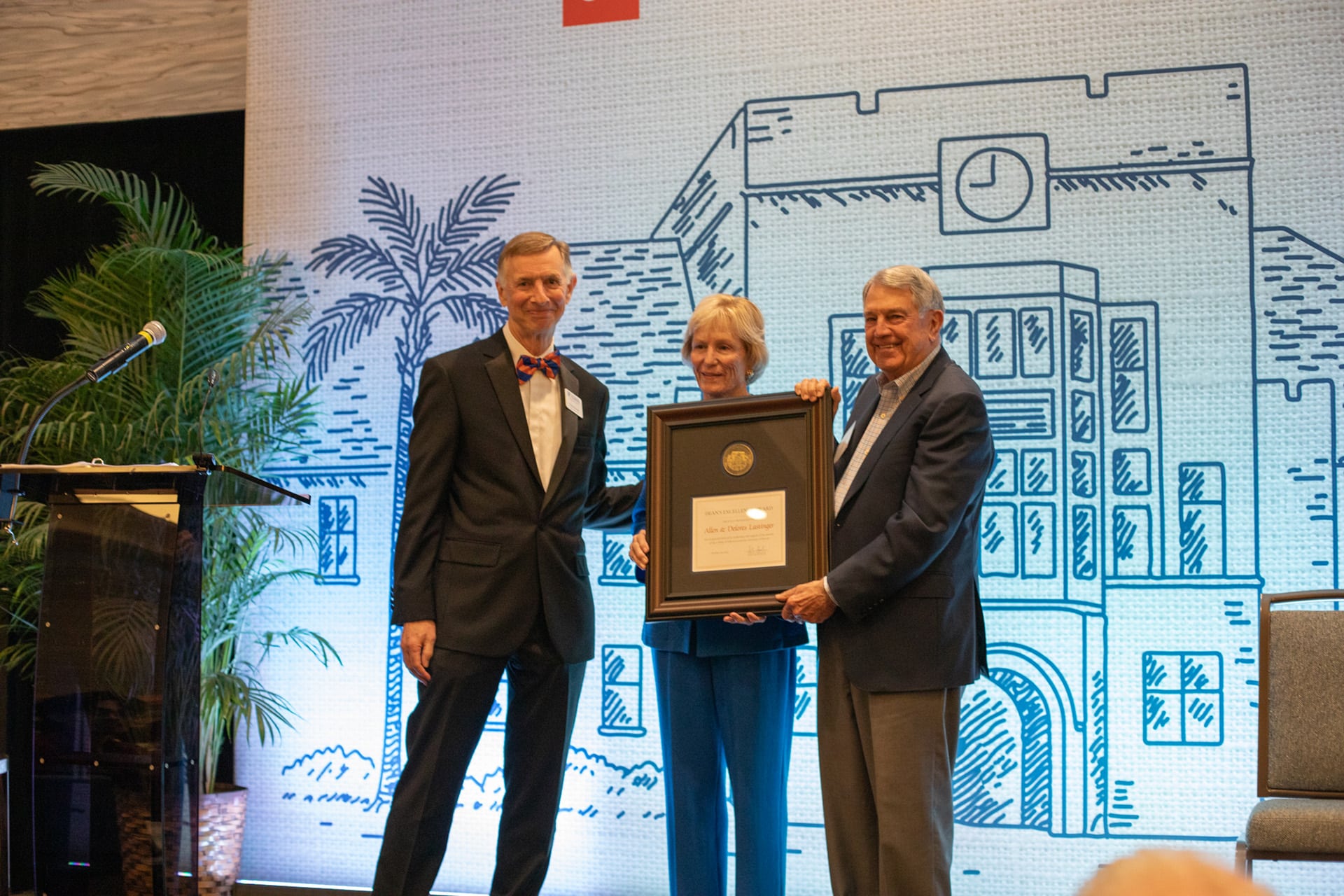 Three people on stage at a UF College of Education event, with two of them holding a large framed award in front of a blue line-art campus backdrop; a podium with a microphone and potted palms are to the left.