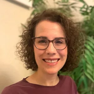 Person with short curly brown hair wearing a maroon T-shirt, indoors with leafy houseplants and warm lighting in the background.