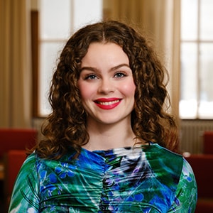 Person with shoulder-length curly brown hair wearing a blue-green patterned top, standing in a softly lit room with tall windows, beige curtains, and red lounge chairs.