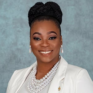 Person in a white blazer over a gray top, wearing layered pearl necklaces and drop pearl earrings, with hair styled in a braided updo, posed against a blue-gray studio backdrop.
