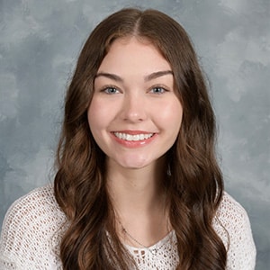 Portrait of a person with long wavy brown hair wearing a white knit sweater against a gray mottled studio backdrop.