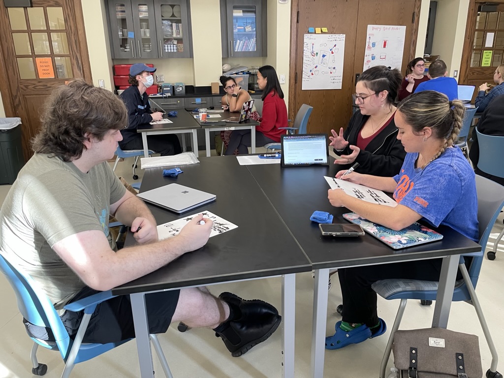 Students collaborate in a classroom at the University of Florida College of Education. Several students are seated around black tables with blue chairs, working on papers and laptops. The room has storage cabinets on the walls and educational posters displayed. One student wears a blue UF shirt, and the setting appears to be a typical UF Teach program working session. Some students are engaged in discussion while others are focused on individual work. The environment suggests an active learning space within an educational setting