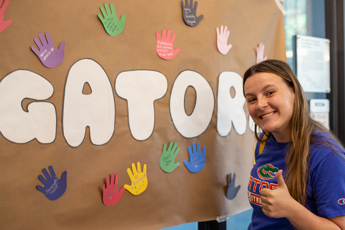 A student giving a thumbs up sign in front of a paper mural that says, "Gator"