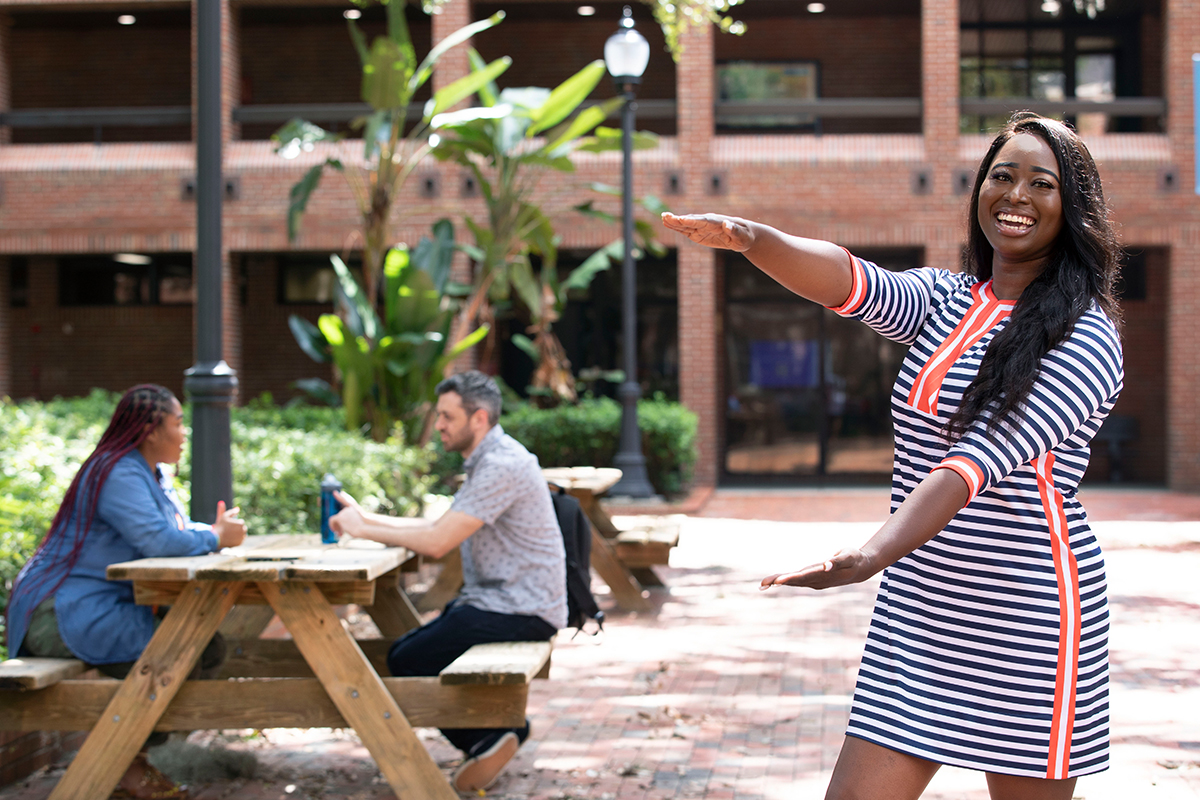 A student doing the Gator Chomp with her hands in the Norman Courtyard while two people sit and chat at a picnic table behind her.